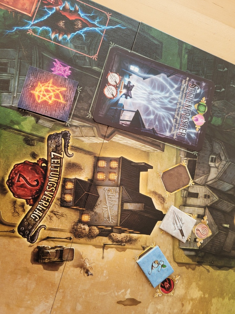 Auf dem Spielplan von "Der Hexer von Salem" ist der Zeitungsverlag zu sehen, daneben die Mauer, hinter der ein Portal sein könnte, und die Monsterkarte Seelenfresser. Außerdem ein freies Gegenstandsfeld sowie ein Feld mit einem Dolch und einem Schauglas.