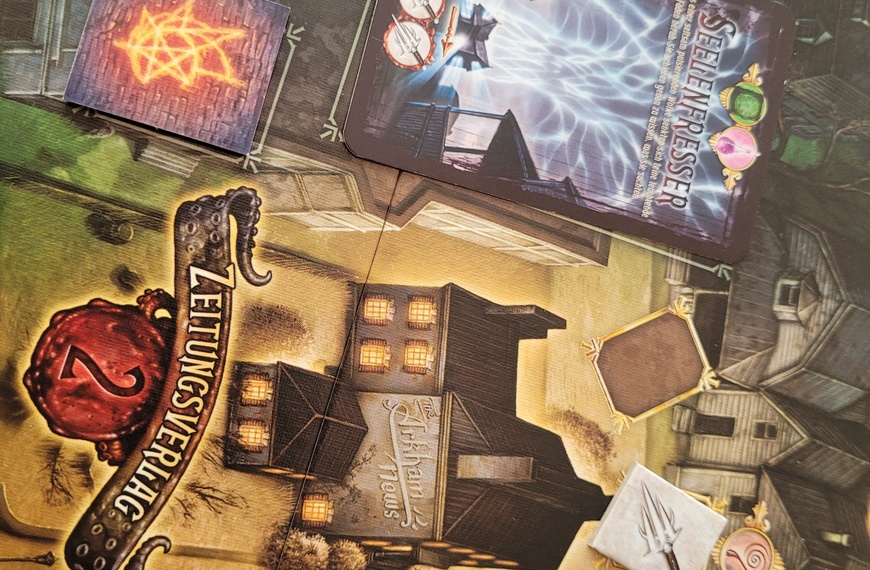 Auf dem Spielplan von "Der Hexer von Salem" ist der Zeitungsverlag zu sehen, daneben die Mauer, hinter der ein Portal sein könnte, und die Monsterkarte Seelenfresser. Außerdem ein freies Gegenstandsfeld sowie ein Feld mit einem Dolch und einem Schauglas.