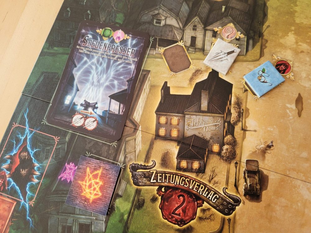 Auf dem Spielplan von "Der Hexer von Salem" ist der Zeitungsverlag zu sehen, daneben die Mauer, hinter der ein Portal sein könnte, und die Monsterkarte Seelenfresser. Außerdem ein freies Gegenstandsfeld sowie ein Feld mit einem Dolch und einem Schauglas.