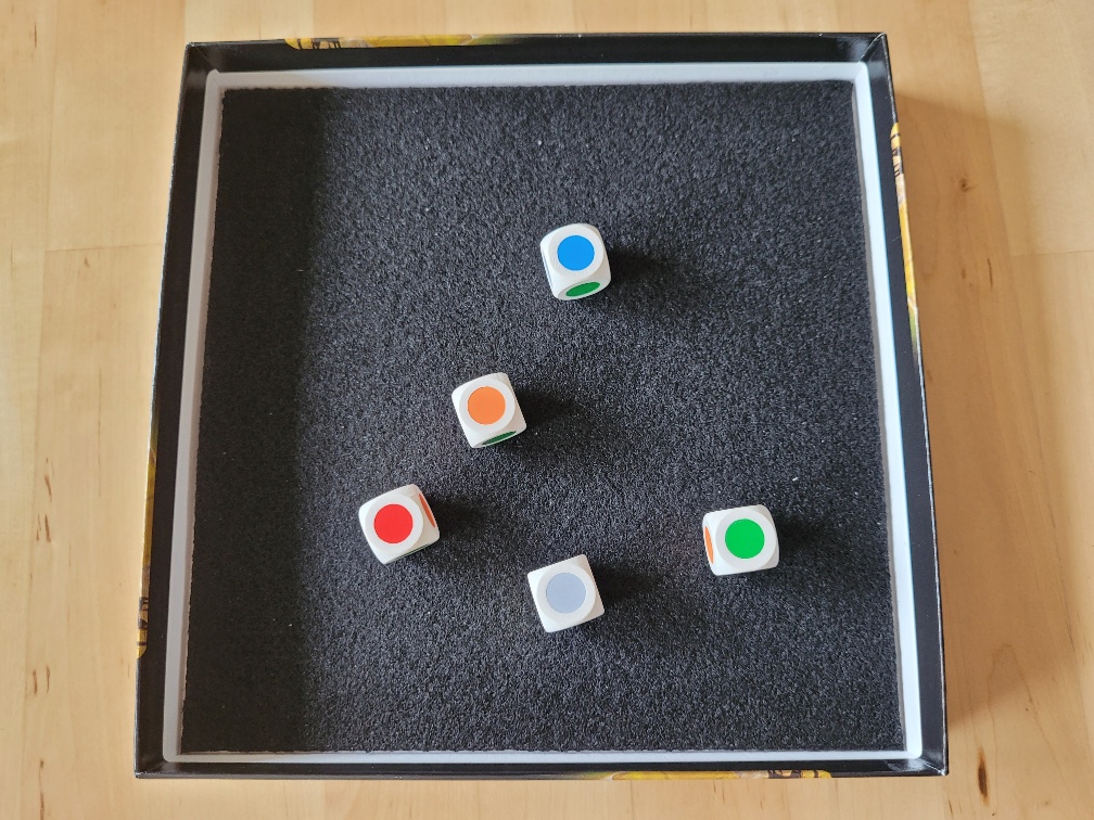 Eine Schachtel mit Stoff im inneren, in der fünf Würfel mit Farbflächen statt Augen liegen.