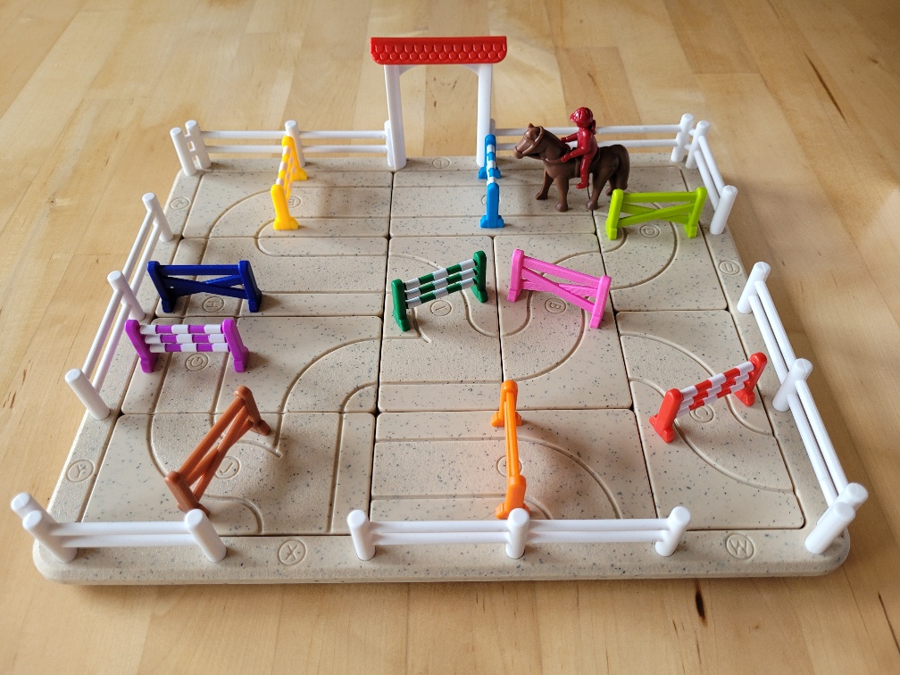 Der dreidimensionale Spielplan von "Schwing die Hufe" sieht aus wie ein Reitplatz mit einem Tor, Zaun und Reiter auf einem Pferd. Dazwischen stehen bunte Hindernisse.