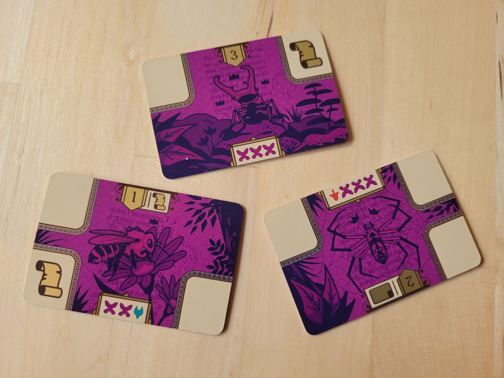 Drei lilafarbene Karten mit einer Spinne einem Hirschkäfer und einer Biene und verschiedenen Symbolen.