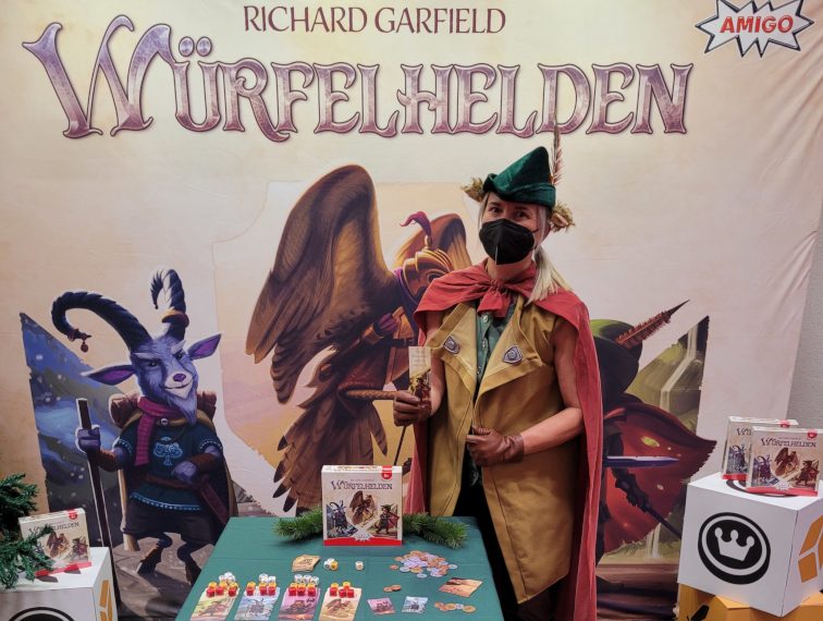 Eine Frau in einer Art Robin-Hood-Kostüm präsentiert auf der Spielemesse 2022 in Essen am Stand das Spiel "Würfelhelden".