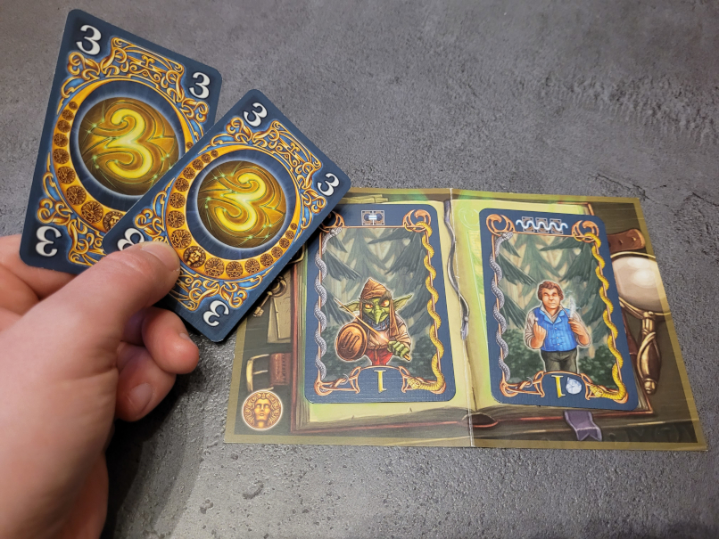 Eine Hand hält zwei Karten mit der Zahl 3. Mit ihnen kann die linke Karte erfüllt werden.