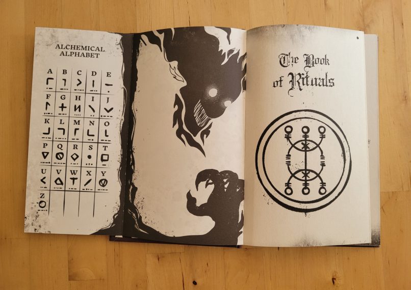 Buchseiten zeigen ein alchemistisches Alphabeth, eine schwarze, unheimliche Figur und den englischen Buchtitel "The Book of Rituals"