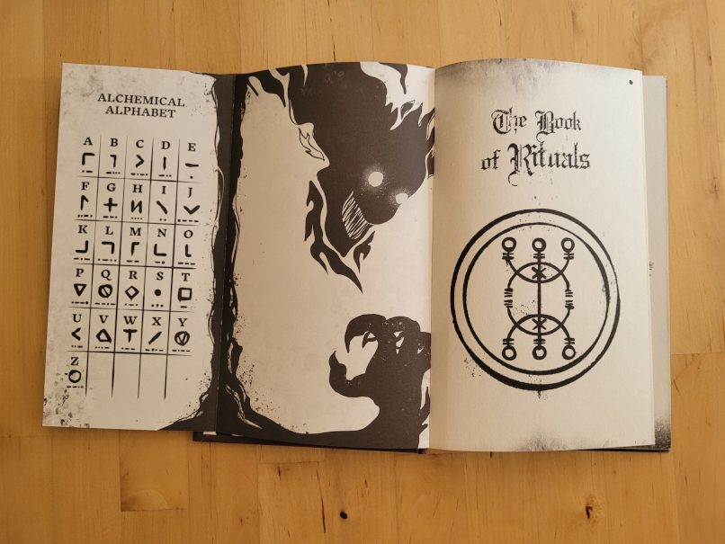 Buchseiten zeigen ein alchemistisches Alphabeth, eine schwarze, unheimliche Figur und den englischen Buchtitel "The Book of Rituals"