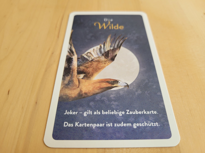 Eine Karte mit einem Greifvogel vor einem Vollmond mit Text "Die Wilde" und "Joker - gilt als beliebige Zauberkarte. Das Kartenpaar ist zudem geschützt".