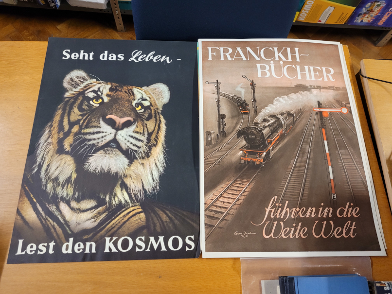 Zwei Plakate werben für Kosmos. Eines zeigt einen Tiger, eines zwei Dampflokomotiven.