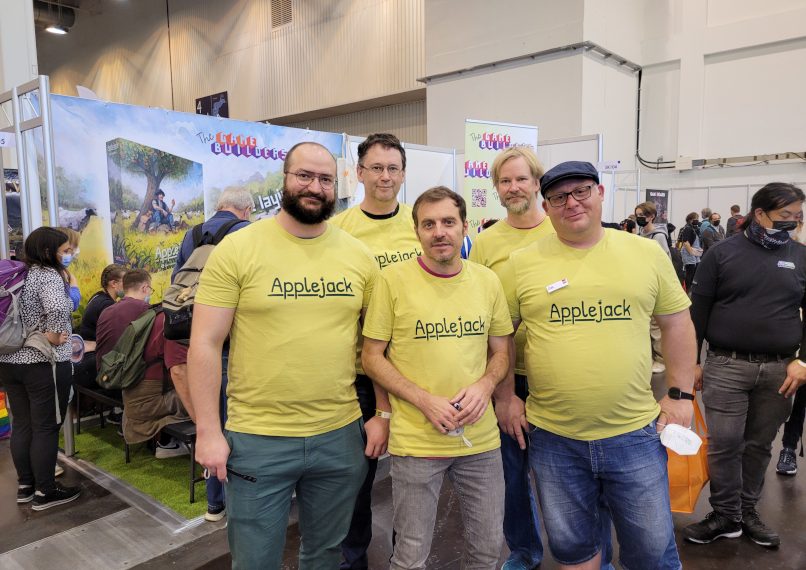 Fünf Männer in gelben T-Shirts mit der Aufschrift Applejack vor dem Messestand von The Game Builders.