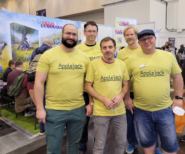 Fünf Männer in gelben T-Shirts mit der Aufschrift Applejack vor dem Messestand von The Game Builders.