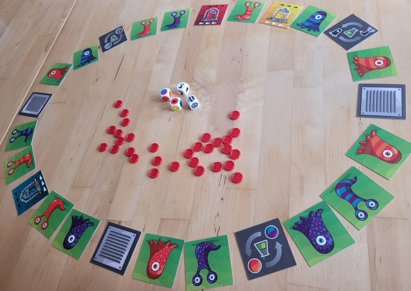 Kärtchen mit Amöben und Aktionskarten liegen in einem Kreis. In der MItte rote Spielchips und vier Würfel.