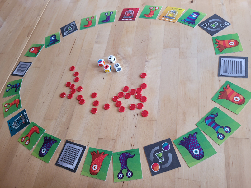 Kärtchen mit Amöben und Aktionskarten liegen in einem Kreis. In der MItte rote Spielchips und vier Würfel.