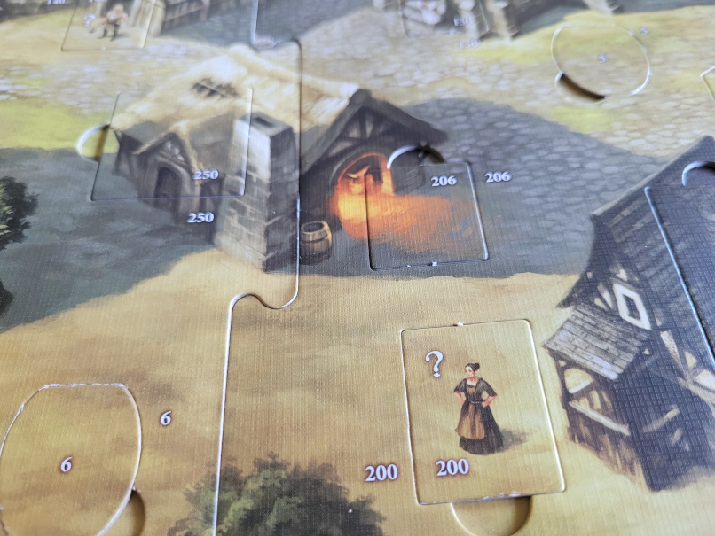 Ein Ausschnitt des Spielplans mit Häusern im Dorf und herausnehmbaren Plättchen, eines davon mit einem Fragezeichen.