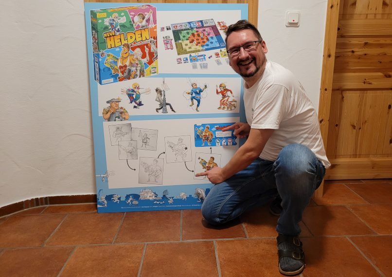 Ein Mann in T-Shirt und Jeans mit Brille kniet vor einem Plakat mit Skizzen und Figuren. Er zeigt mit den Händen auf das Plakat.