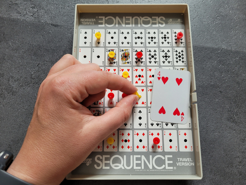 Eine Hand steckt einen gelben Marker an die Stelle der Herz-Vier im Spielplan von "Sequence Trafel". Daneben liegt die Karte Herz-Vier.