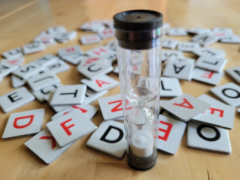 Eine Sanduhr und dahinter Plättchen mit roten und schwarzen Buchstaben - das Spielmaterial von "Wörterklauer".