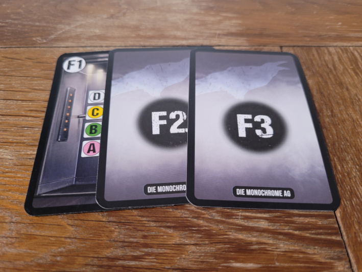 Eine Karte, die einen halbe verdeckten Fahrstuhl zeigt und zwei Karten, auf deren Rückseite "F2" und "F3" steht.