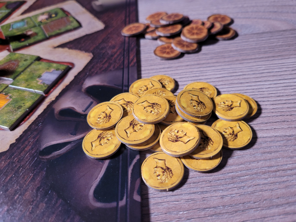 Ein Stapel Münzen mit einem Caesaren-Kopf und ein Stapel Brote liegen neben dem Spielplan von "Carpe Diem".