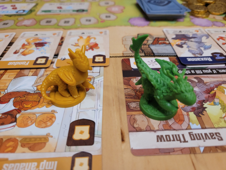 Ein gelber und ein grüner Drachen stehen auf zwei Läden. Im Hintergrund ist weiteres Spielmaterial zu sehen.