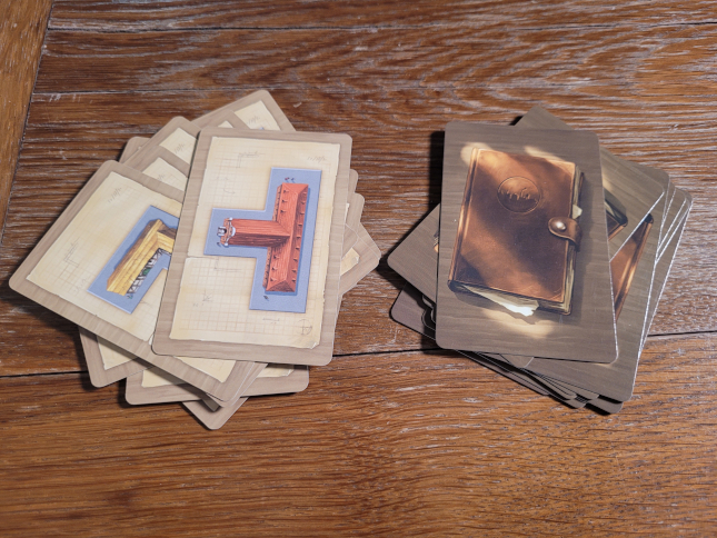 Zwei Kartenstapel, einer zeigt tetrisförmige Bauteile, einer eine Lederkladde.