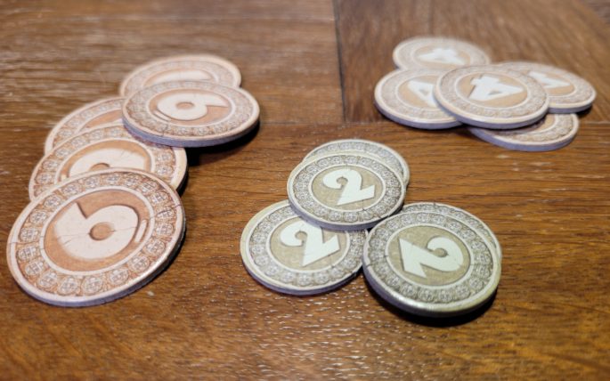 Göttersteine sehen aus wie Münzen mit den Werten zwei, vier und sechs. Sie liegen auf einem Tisch.