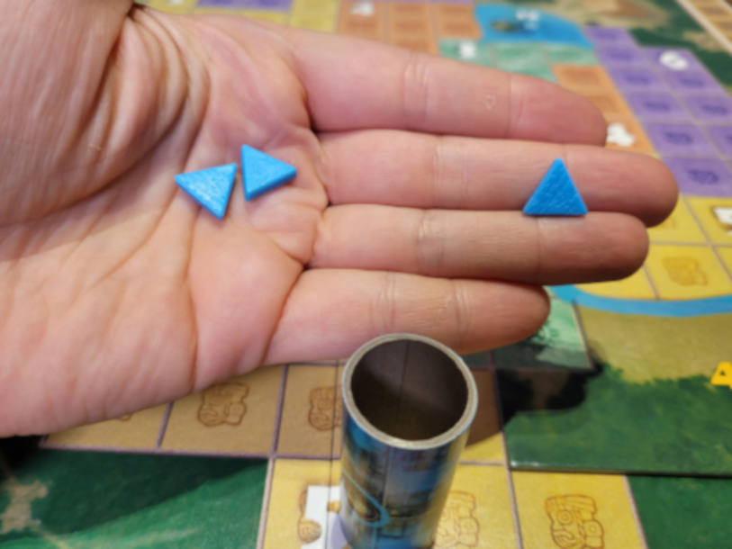 Eine Hand hält drei dreieckige, blaue Energiesteine über dem Spielplan von "Pacal's Rocket".