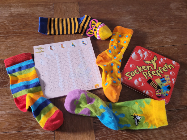 Das Spielmaterial von Socken pfeffern: Vier bunte Socken und ein Wertungsblock neben dem Dosendeckel.