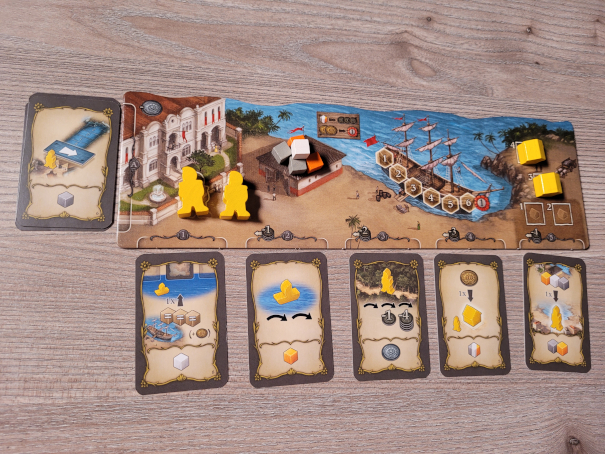 Ein Spielertableau aus "Valbaraiso" mit Figuren, Waren und Karten. Ein Schiff in einer Bucht und ein großes Haus sind zu sehen.