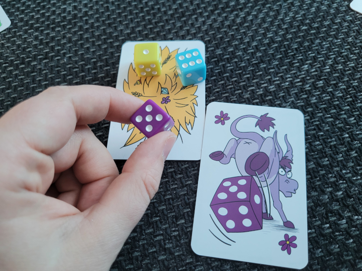 Eine Hand hält den lilafarbenen Würfel über der Heuhaufen-Karte, auf der schon ein gelber und ein hellblauer Würfel liegen. Daneben liegt die lilafarbene Wütender-Esel-Karte.