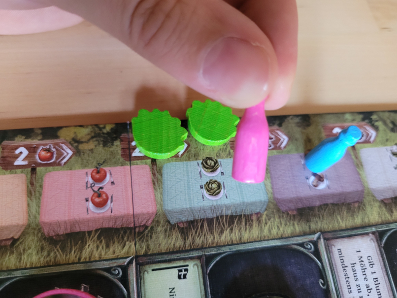Eine Hand hält die pinke Milchflaschen-Figur über einem Tisch auf dem Spielplan von "Reykholt".