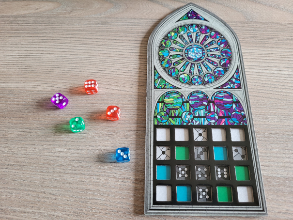 Neben einem Spielertableau in Kirchenfensterform liegen fünf Würfel in verschiedenen Farben.