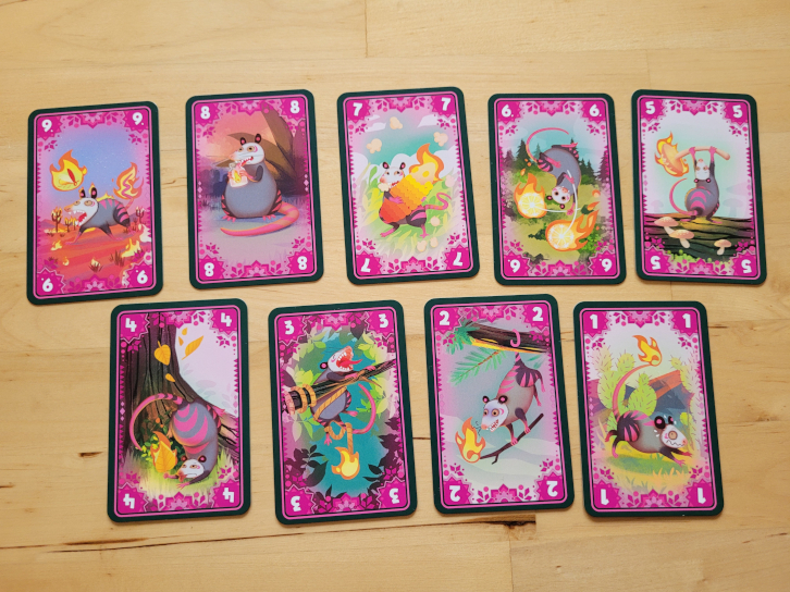 Neun Karten mit pinkfarbenem Rand. Die Karten haben die Werte Eins bis Neun und zeigen je ein Opossum.
