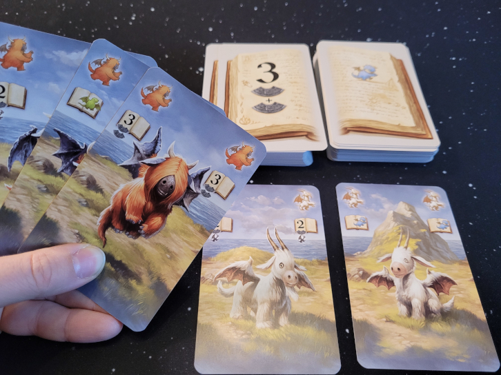 Eine Hand hält drei Karten über zwei Kartenstapel und zwei ausliegende Karten.