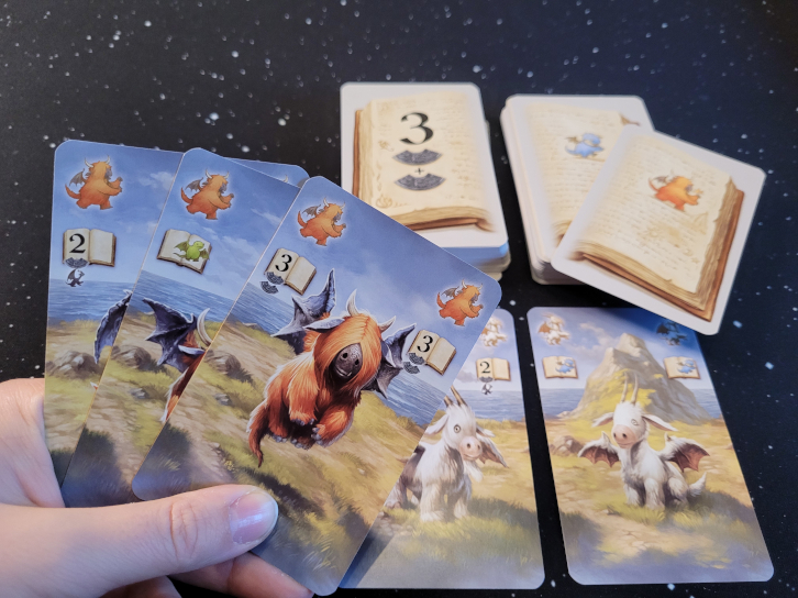 Eine Hand hält drei Karten über zwei Kartenstapel und zwei ausliegende Karten. Auf dem rechten Stapel liegt eine Karte, die den Stapel verändert.