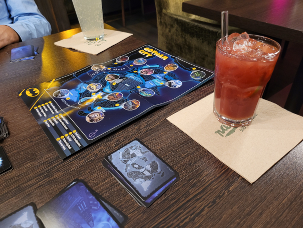 Ein Tisch mit einem Spielplan, einigen Karten und einem Glas mit Eiswürfeln und rotem Getränk.