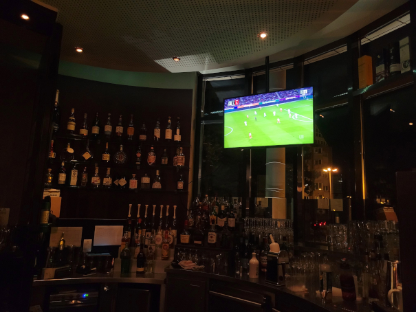 Eine Bar mit Flaschen und Gläsern, auf einem TV-Bildschirm läuft ein Fußballspiel.