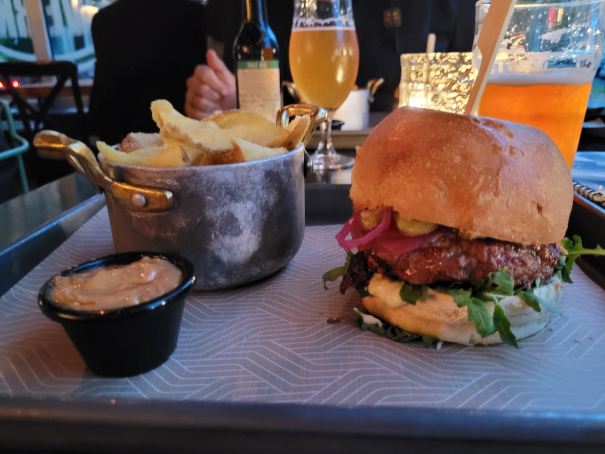 Ein Burger auf einem Tablett mit Pommes und Dipp, dahinter zwei Biergläser.