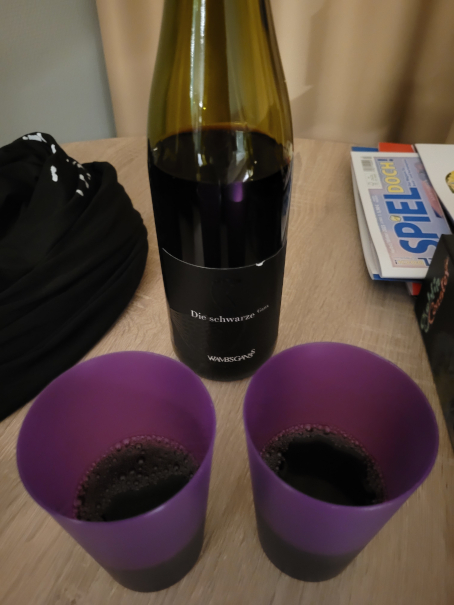 Eine Flasche Rotwein, davor zwei lilafarbene Becher mit Rotwein.
