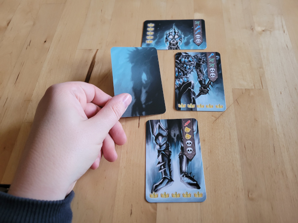 Eine Hand hält die vierte, noch verdeckte Karte, an bereits drei aufgedeckte Karten, die zusammengesetzt den Skelettkönig ergeben.