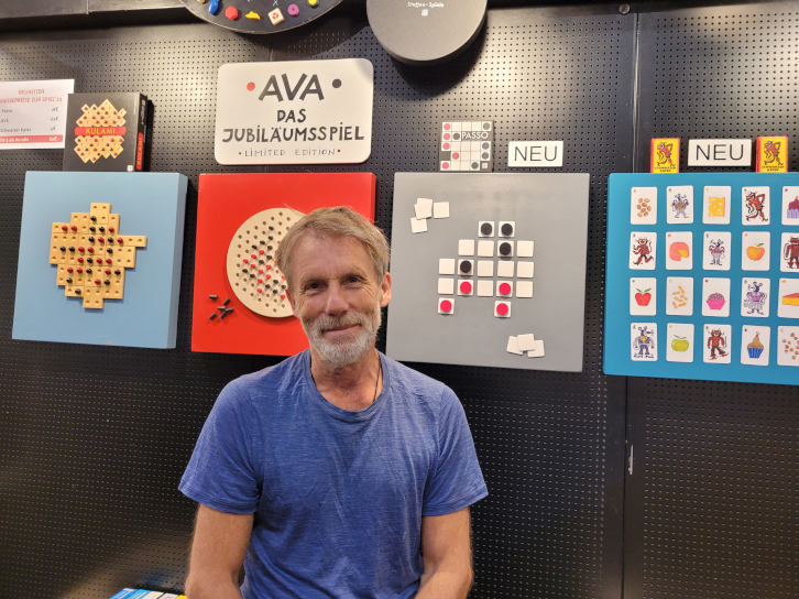 Ein Mann in blauem T-Shirt steht vor einer Wand, an der Spiele wie Bilder aufgehängt sind.