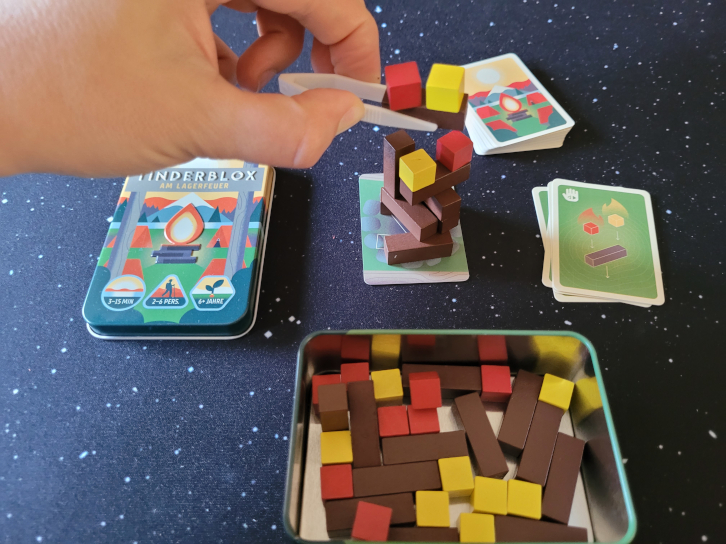 Das Spielmaterial von Tinderblox. In der Mitte ein Stapel aus den Holzbauteilen. Eine Hand setzt mit einer Pinzette drei gestapelte Bauteile darauf.