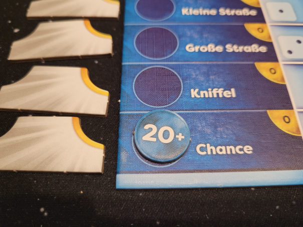 Ein rundes Plättchen mit "20 +" liegt auf einem Spielplan neben dem Wort "Chance".