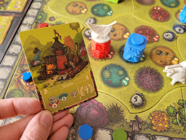 Eine Hand hält eine Karte neben zwei Gebäuden auf dem Spielplan von "Kleine Völker, großer Garten".