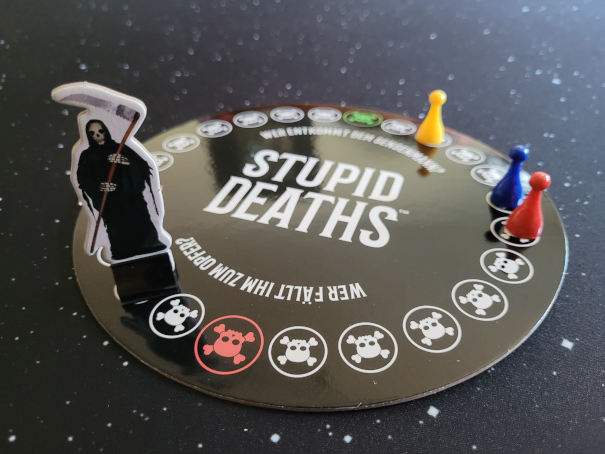 Der Spielplan von "Stupid Deaths" mit Sensemannfigur und drei Kunststoffspielfiguren in Blau, Rot und Gelb.