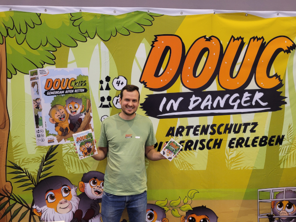 Ein Mann hält das Spiel "Douc in Danger" vor einer Werbewand, auf der ebenfalls der Name des Spiels steht.