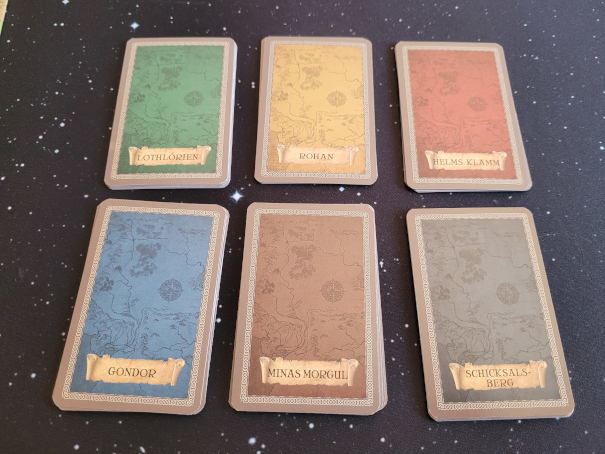 Sechs Kartenstapel in Grün (Lothlorien), Gelb (Rohan), Rot (Helms Klamm), Blau (Gondor), Braun (Minas Morgul) und Schwarz (Schicksalsberg).