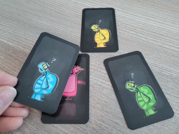 Eine Hand hält eine Karte mit einer Comic-Schildkröte über drei Kartenstapel aus "Dodelido".