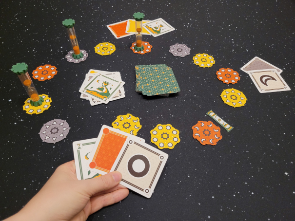 Eine Hand hält drei Karten, dahinter ist das Spielmaterial von "Quicksand" zu sehen.