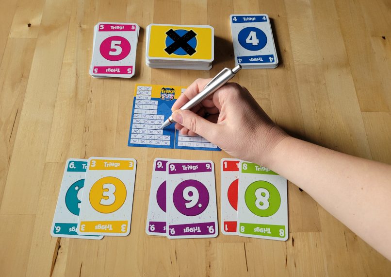 Das Spielmaterial von "Triggs": Karten mit Zahlen und ein Wertungsblatt. Eine Hand hält über das Blatt einen Stift.