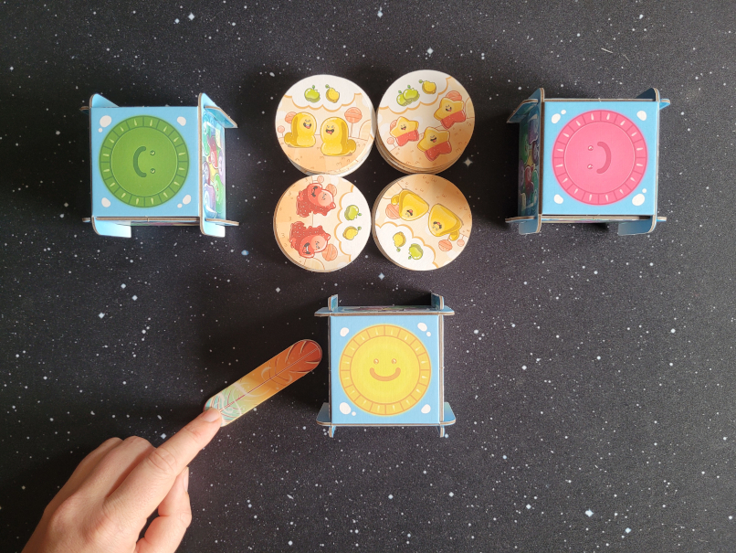Eine Hand hält eine Pappfeder neben eine Kartonbox. Daneben liegt weiteres Spielmaterial.